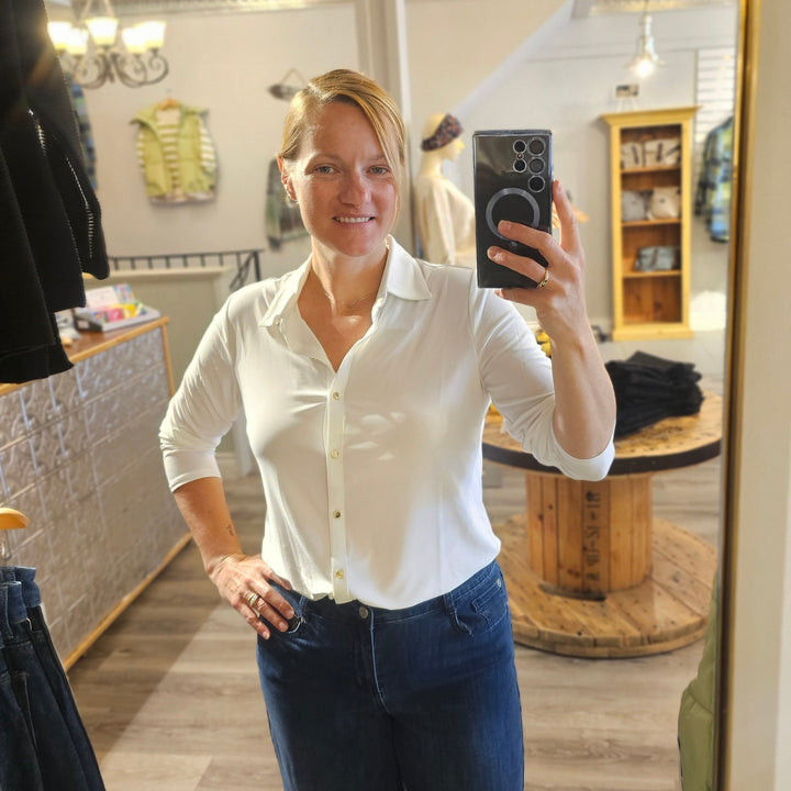 Person taking a mirror selfie wearing a white shirt and blue jeans in a room with a wooden cabinet.