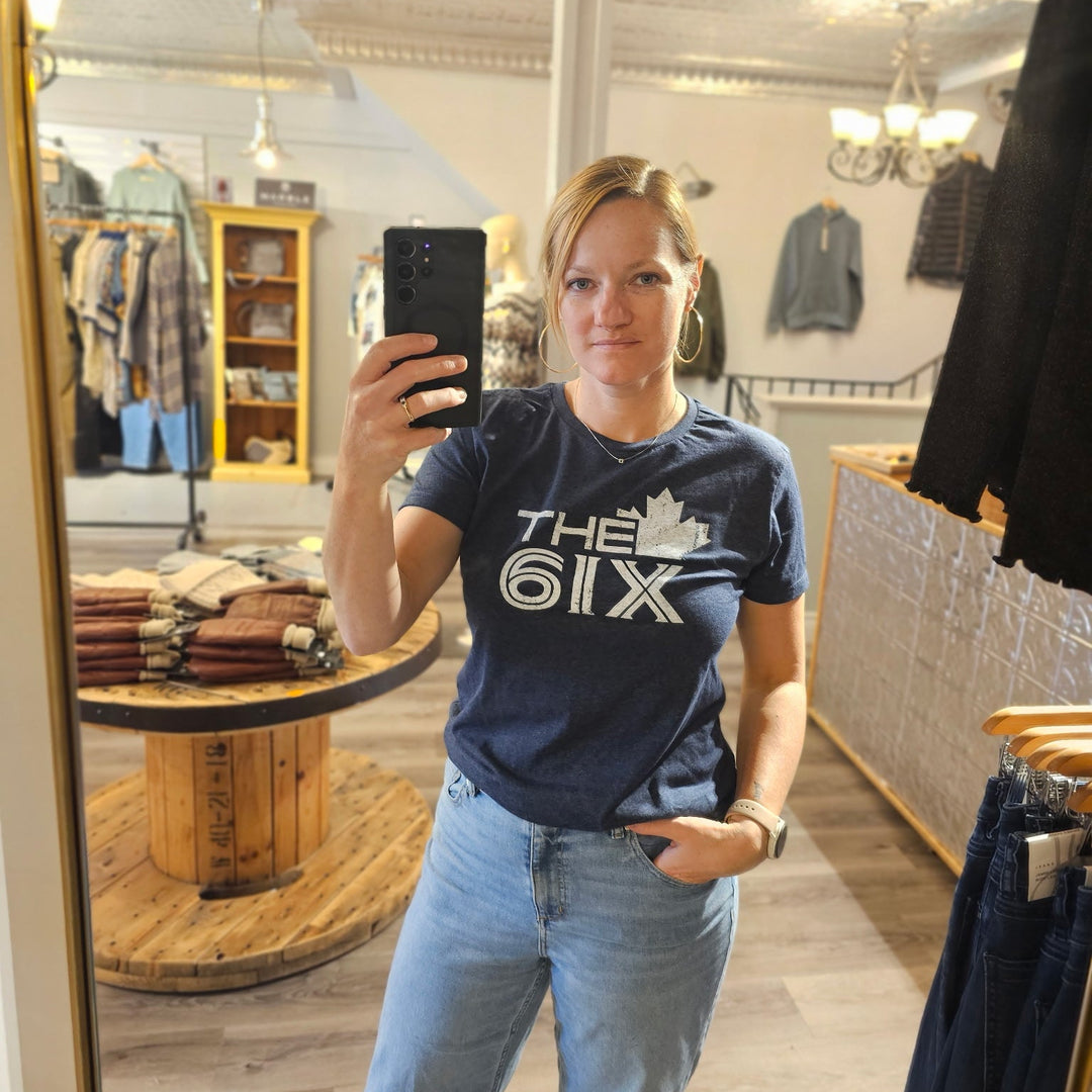 Person taking a mirror selfie wearing a blue t-shirt with 'The 61x' logo in a store.