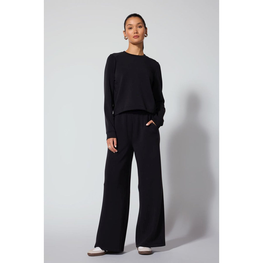 A model stands against a light gray studio background wearing a black long-sleeve cropped top and matching black wide-leg trousers, paired with white sneakers.