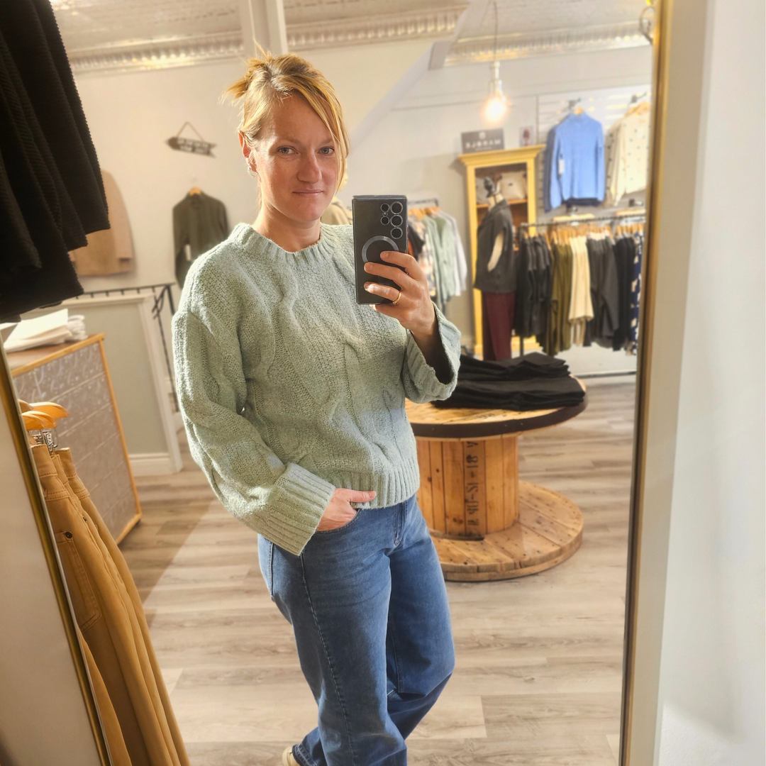 Person taking a mirror selfie wearing a green sweater and blue jeans in a clothing store.